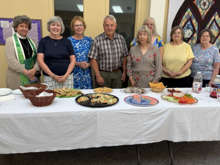 Seven adults stand behind a long table laden with cookies, sandwiches, fruit, and drinks at a community gathering. A colorful quilt hangs on the wall behind them.
